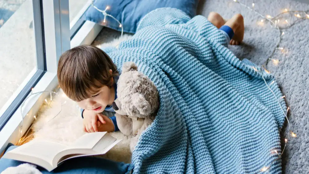 Junge liegt gemütlich in eine Decke eingekuschelt auf dem Boden und liest seinem Teddybär vor