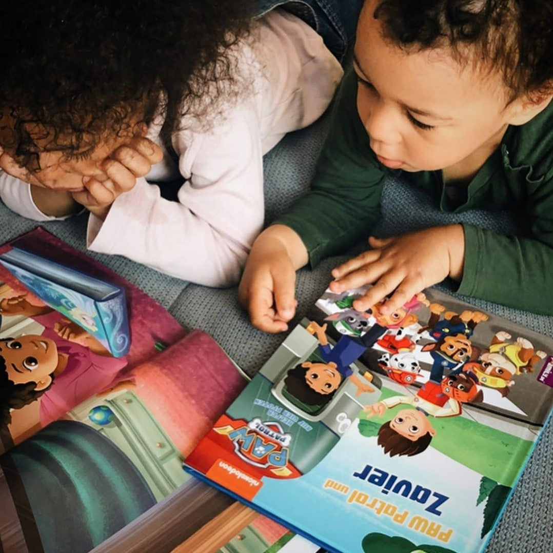 Zwei kleine Kinder liegen auf einem Bett und lesen farbenfrohe Bilderbücher, darunter eines mit den Figuren der Paw Patrol. Sie sehen konzentriert aus und beschäftigen sich mit den Geschichten.