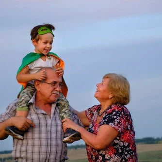 Großvater hat Kind mit Superhelden-Kostüm auf den Schultern und Großmutter lächelt ihm zu