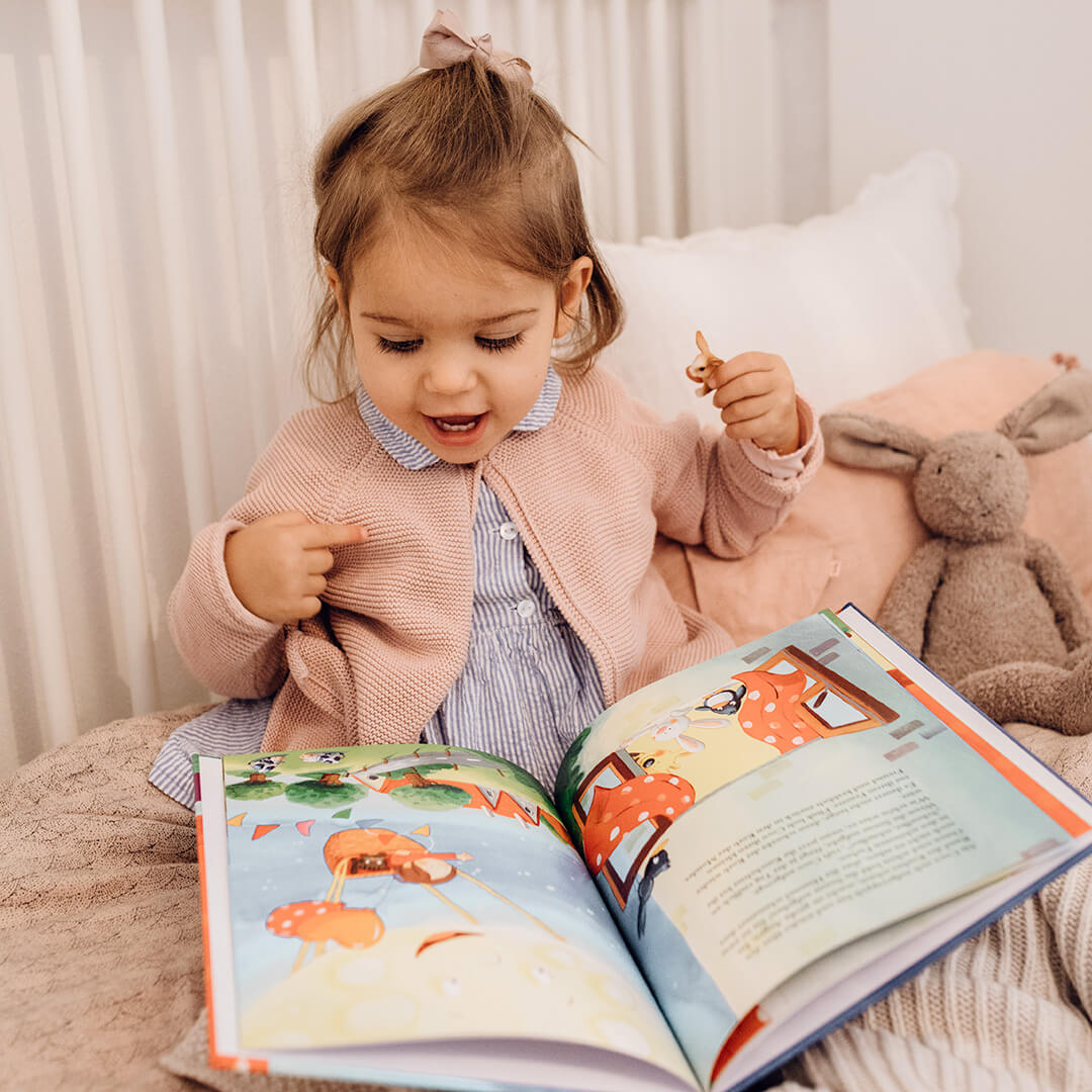 Ein kleines Kind sitzt auf einem Bett, lächelt und zeigt auf ein aufgeschlagenes, buntes Bilderbuch, neben dem ein Plüschhase liegt. Es trägt eine rosafarbene Strickjacke und ein blaues Kleid und sieht aus, als sei es in seinem gemütlichen Schlafzimmer mit der Geschichte beschäftigt.