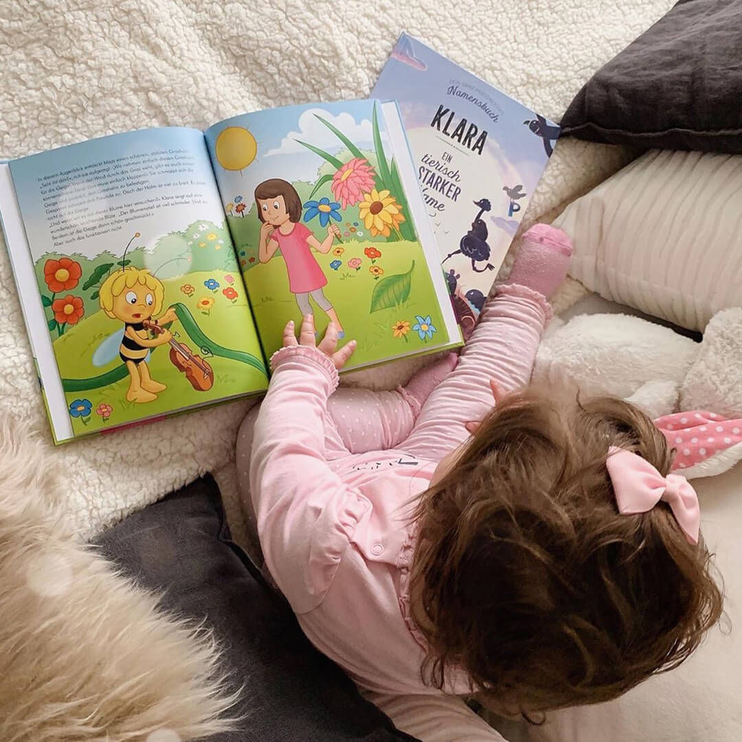 Ein kleines Kind mit einer rosa Schleife im Haar sitzt auf einem Bett und betrachtet ein aufgeschlagenes Bilderbuch mit bunten Blumen und einem Mädchen. Ein anderes Buch mit dem Titel Klara liegt in der Nähe.