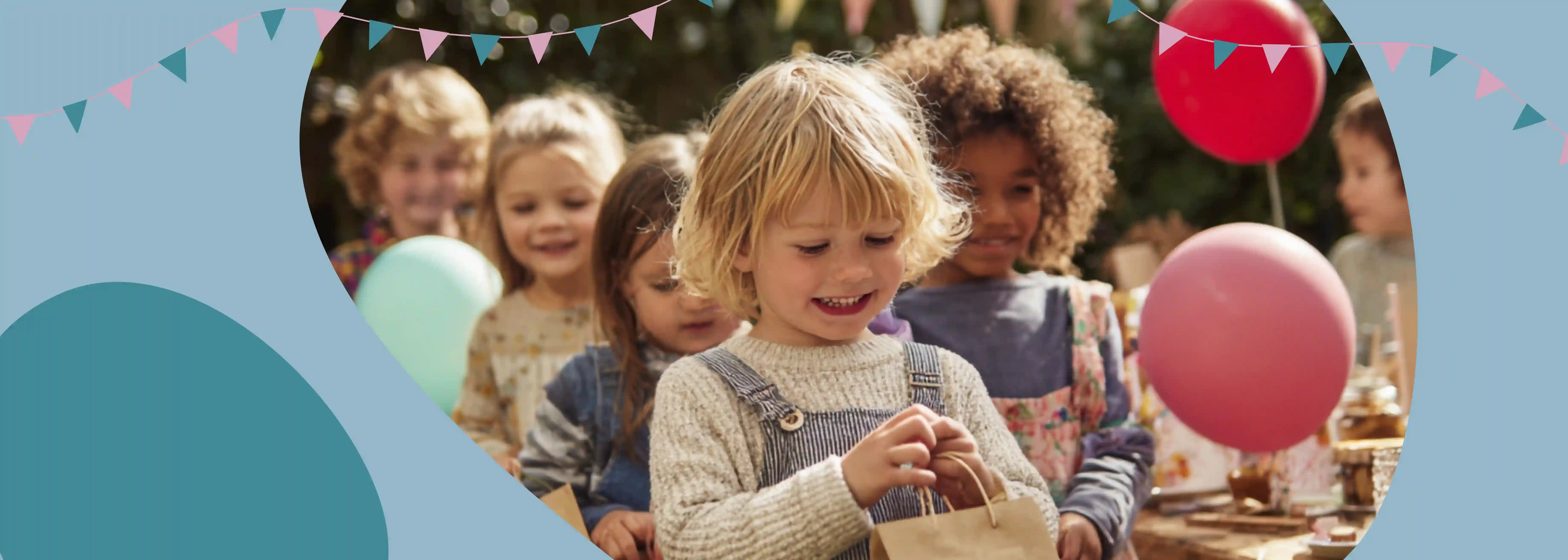 Kind hält lächelnd eine Mitgebsel-Tüte in der Hand, hinter ihm stehen weitere Kinder. Im Hintergrund ist ein Geburtstagstisch und eine Girlande zu sehen.