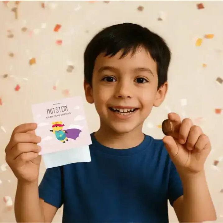 Junge hält Mutstein hoch mit breitem Lächeln im Gesicht. Im Hintergrund ist goldenes Konfetti zu sehen.