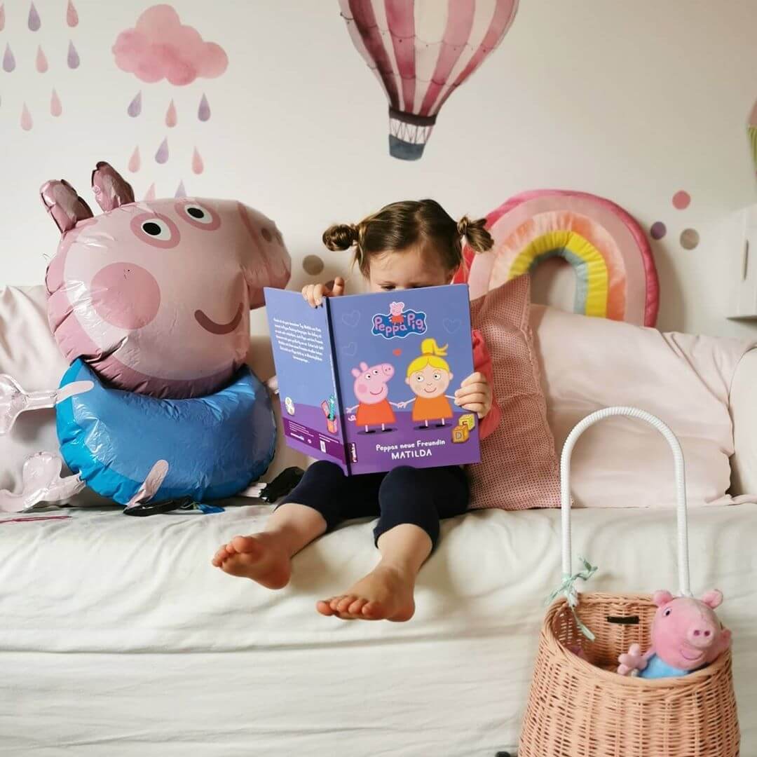 Ein kleines Kind sitzt auf einem Sofa und liest ein Peppa Pig-Buch, umgeben von Dekorationen mit Peppa Pig-Motiven, darunter ein Ballon und ein Spielzeug, in einem Raum mit pastellfarbener Wandkunst und einem rosa Korb in der Nähe.