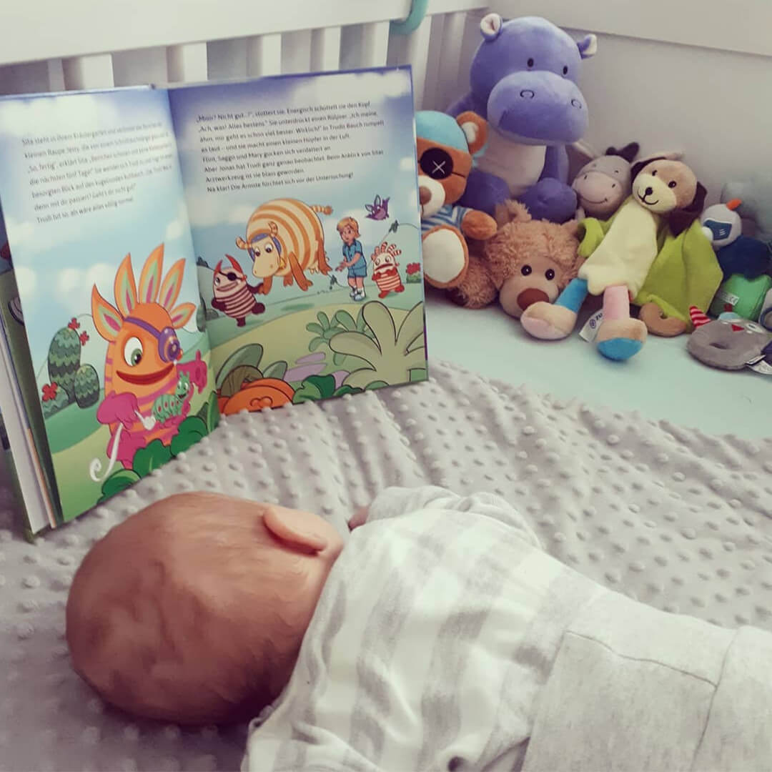 Ein Baby liegt auf einer grauen Decke vor einem aufgeschlagenen Bilderbuch, während im Hintergrund eine Reihe von bunten Plüschtieren an der Wand aufgereiht ist.
