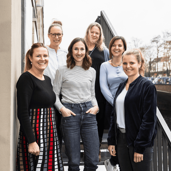 Sechs Frauen stehen gemeinsam auf einer Außentreppe und lächeln in die Kamera. Gekleidet in Freizeit- und Business-Casual-Outfits posieren sie selbstbewusst vor einem Gebäude mit buntem Herbstlaub und Bäumen im Hintergrund.