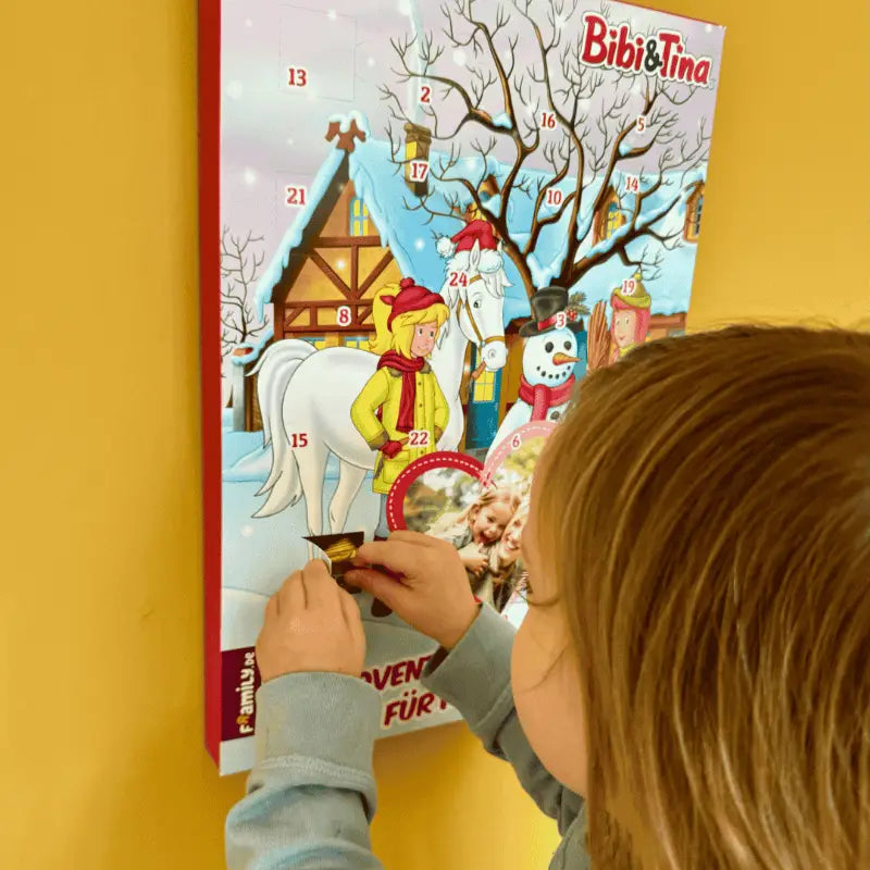 Ein Kind öffnet aufgeregt ein Türchen des Framily Bibi & Tina Adventskalenders, der an einer gelben Wand hängt.