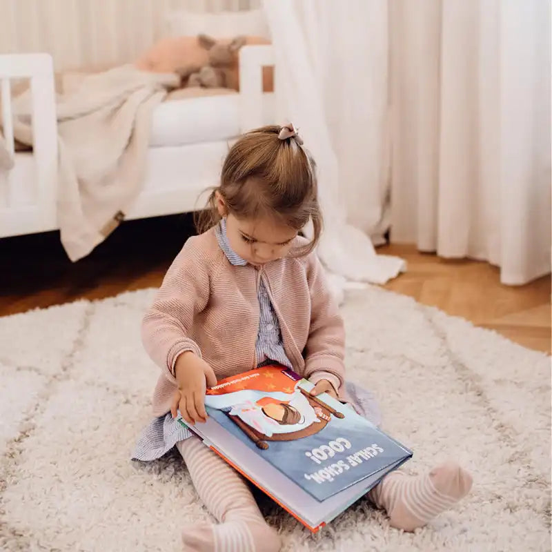 Ein junges Mädchen mit hellbraunem Haar in einem Pferdeschwanz sitzt auf einem weichen Teppich und liest "Deine Gute-Nacht-Geschichte" von Framily. Sie trägt eine rosa Strickjacke, eine gestreifte Strumpfhose und ein Kleid. Im Hintergrund sind ein weißes Bett und beigefarbene Vorhänge zu sehen.