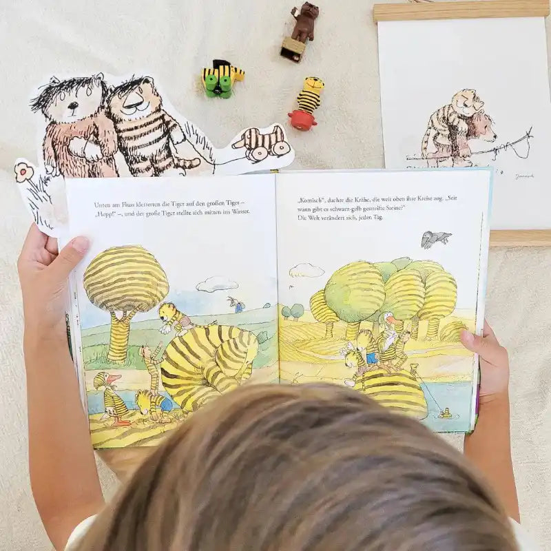 Ein Kind hält "Janosch - Tiger & Bär und du" von Framily aufgeschlagen in der Hand, ein bunt illustriertes Buch. Auf dem Tisch neben dem aufgeschlagenen Buch liegen Papptigerausschnitte und passendes Spielzeug.