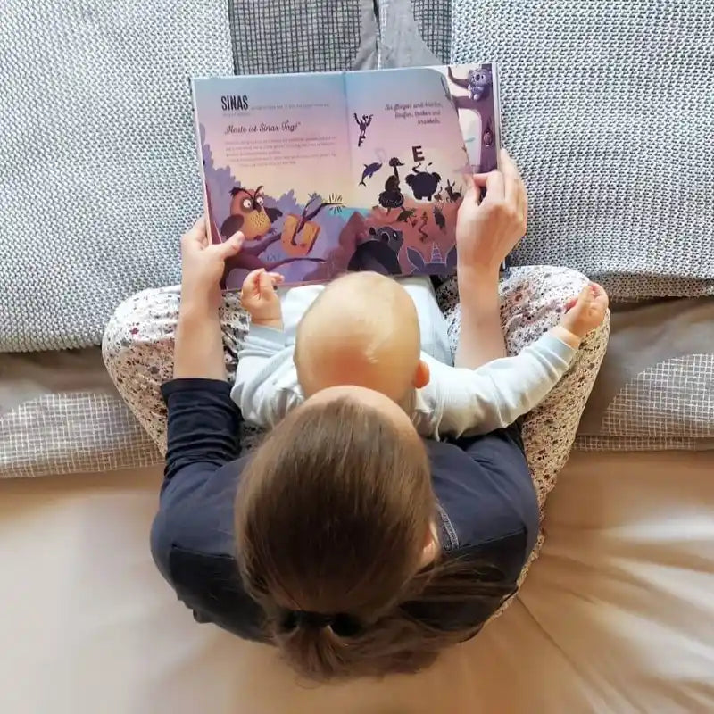 Eine Frau sitzt mit einem Baby auf dem Schoß auf einem Bett und liest das farbenfrohe „Dein Namensbuch“ von Framily vor. Von oben betrachtet, hebt das Bild das offene Buch und die gemütliche Atmosphäre hervor.