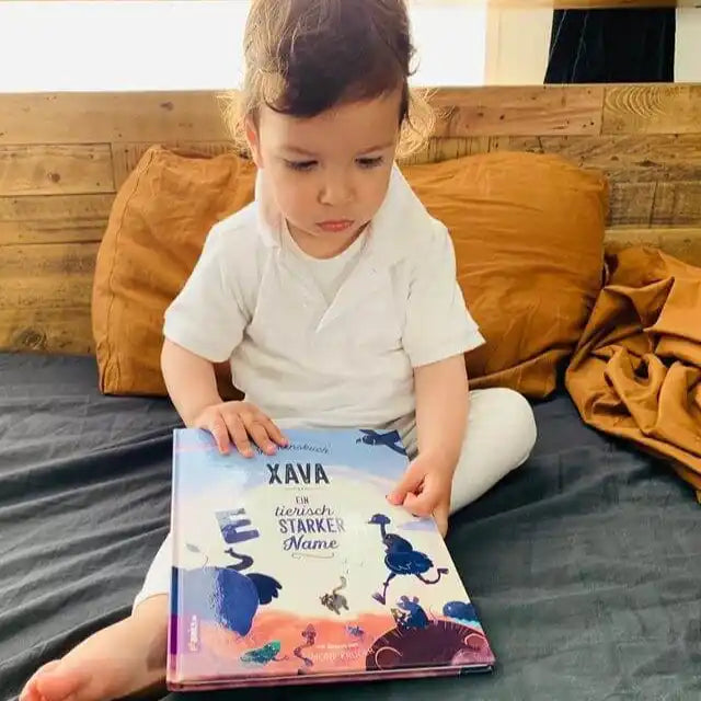 Ein Kleinkind in einem weißen Outfit sitzt auf einem Bett mit senffarbenen Kissen, blickt nach unten und berührt den Einband von Framilys „Dein Namensbuch“.