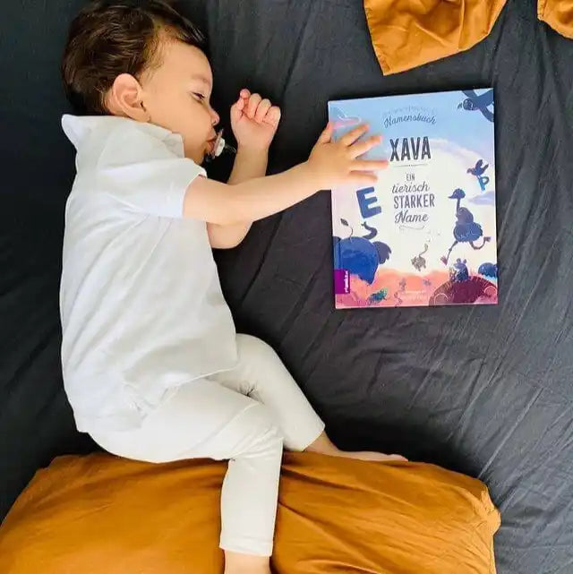 Ein kleines, weiß gekleidetes Kind schläft auf einem dunklen Bett mit einem senfgelben Kissen und berührt sanft den Einband von „Dein Namensbuch“ von Framily.