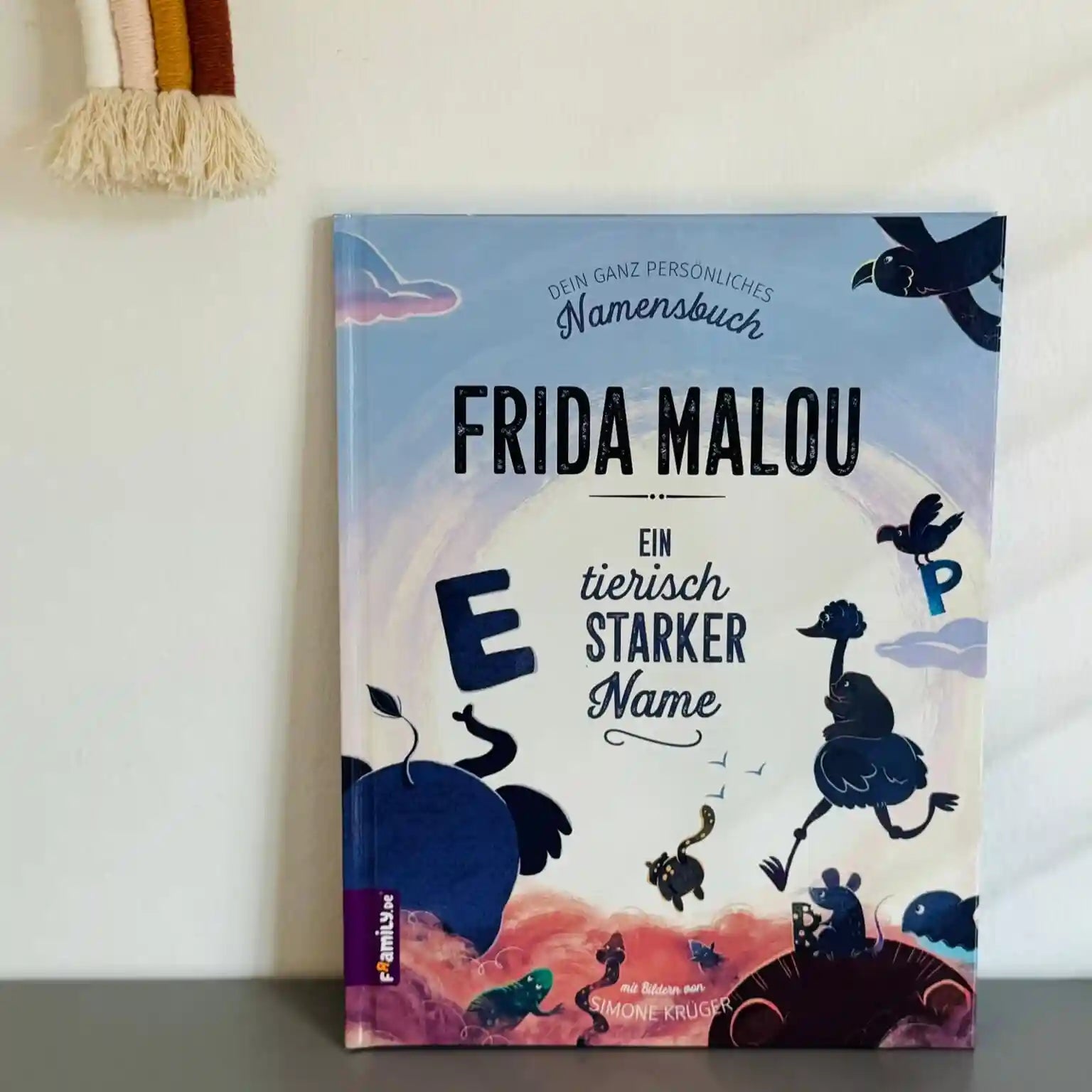 Das Framily Namensbuch steht vor einer weißen Wand. Das Cover zeigt verspielte Tier-ABC-Illustrationen, bunte Buchstaben und einen pastellfarbenen Himmel.