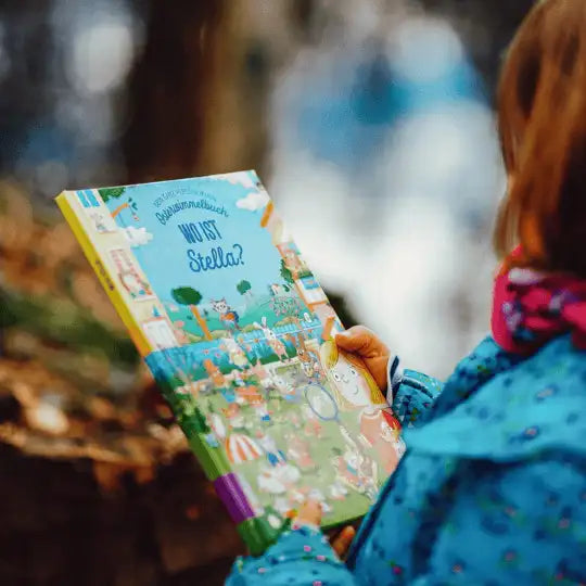 Ein Kind in einer blauen Jacke hält das Osterwimmelbuch von Framily mit einer personalisierten Osterreise in der Hand. Es ist draußen im Freien unterwegs.