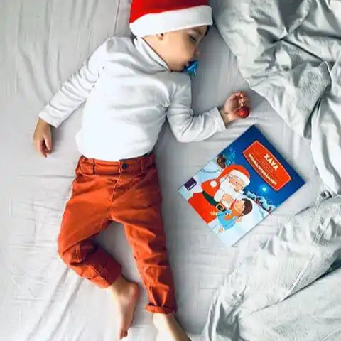 Ein kleines Kind mit Weihnachtsmannmütze, weißem Hemd und orangefarbener Hose schläft neben "Dein Weihnachtsmärchen" von Framily und hält ein kleines rotes Ornament.