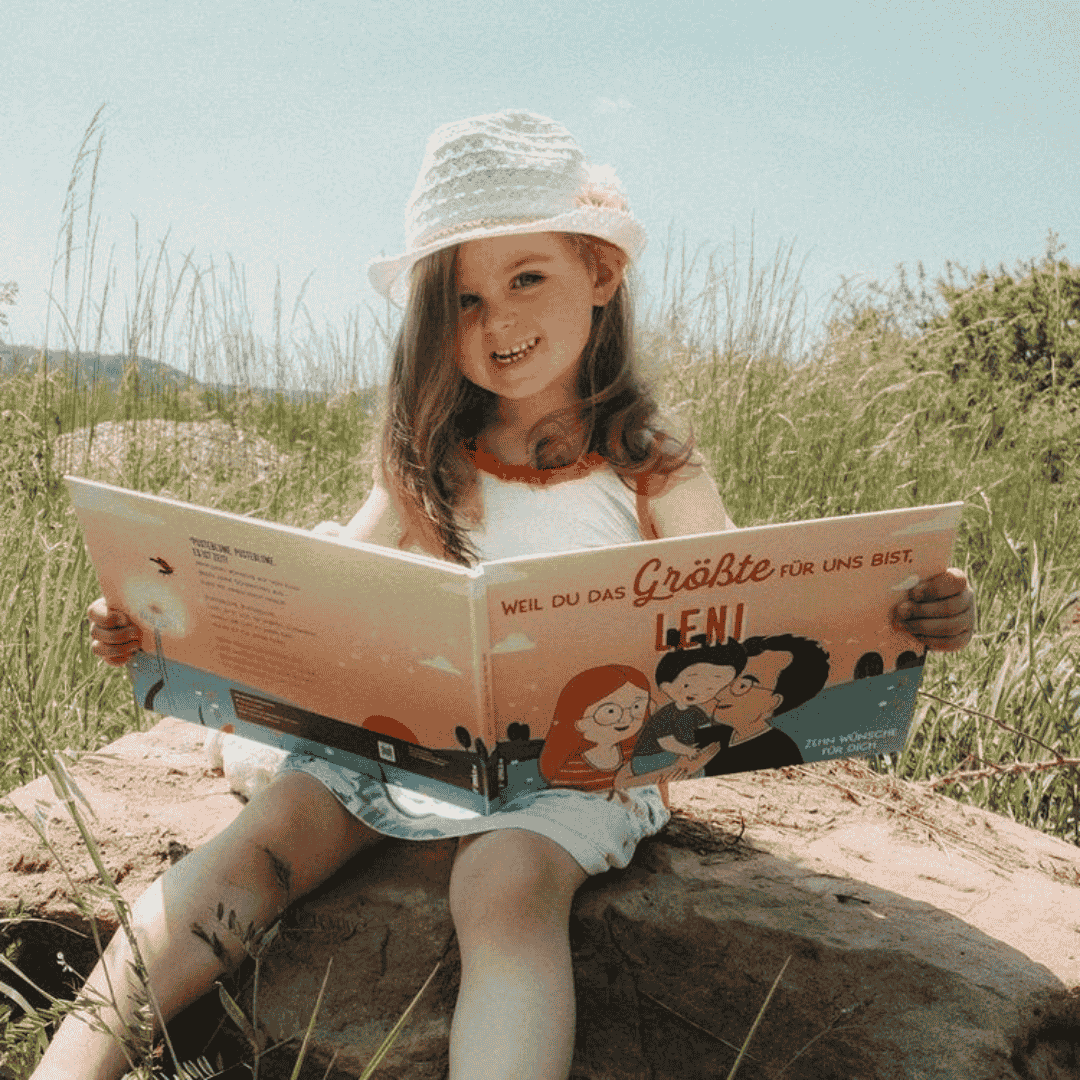 Ein junges Mädchen mit weißem Hut und Sommerkleid sitzt auf einem Felsen im Freien und hält lächelnd ein großes illustriertes Märchenbuch mit dem Titel Leni in der Hand, in dem sie liest. Die Szene ist hell, grasbewachsen und einladend.