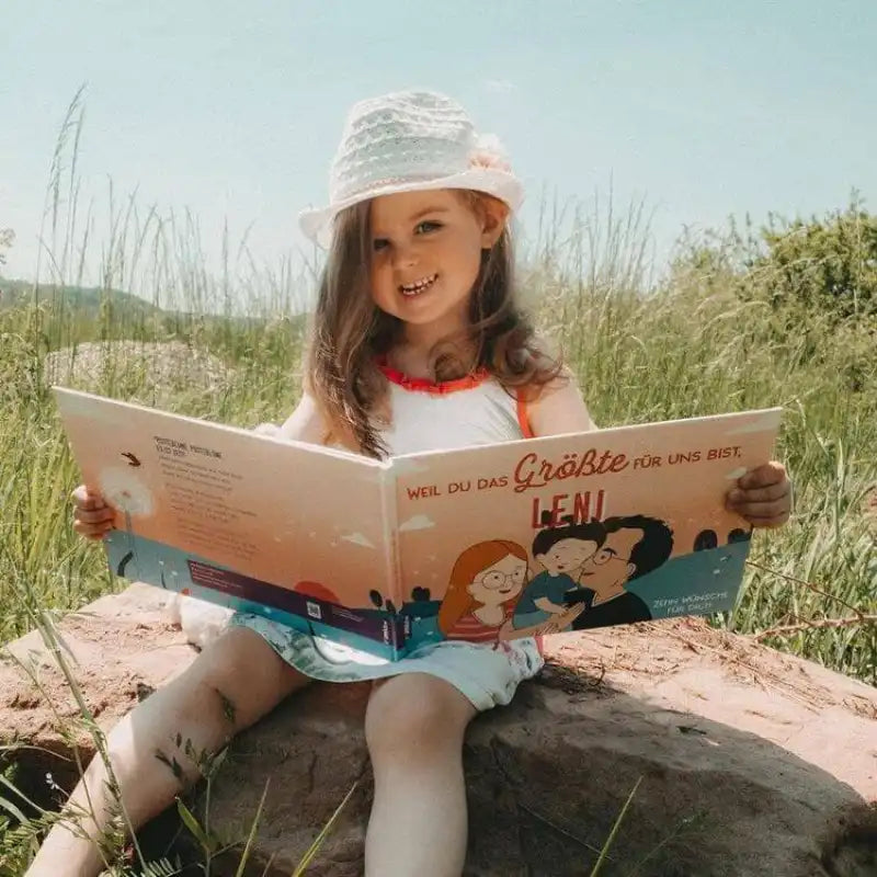 Ein junges Mädchen in einem weißen Kleid und Sonnenhut sitzt auf einem Felsen im Freien und hält lächelnd "Weil du das Größte für mich bist" von Framily in der Hand, umgeben von grasbewachsenen Feldern.
