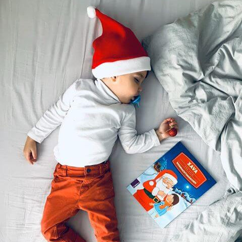 Ein kleines Kind mit roter Weihnachtsmannmütze, weißem Hemd und roter Hose schläft auf einem Bett neben einem weihnachtlichen Buch und einem festlichen roten Ornament.