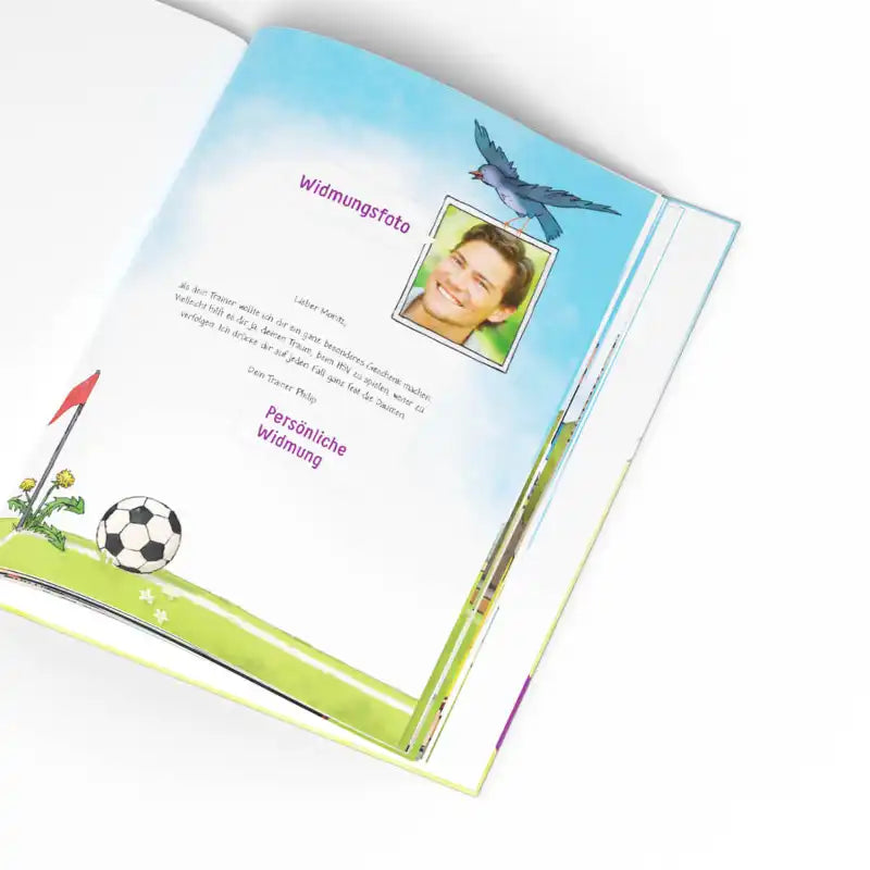 Das aufgeschlagene Kinderbuch "Dein großes Fußballspiel" von Framily zeigt eine bunte Seite mit einem Fußball auf dem Spielfeld, Gras, Blumen, einem Vogel, einem lächelnden Portraitfoto, deutschem Text und Platz für eine persönliche Widmung.