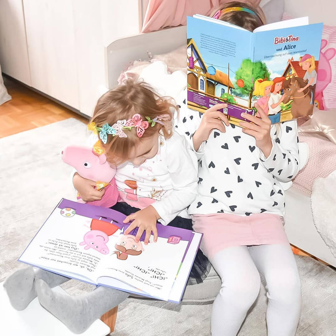 Zwei junge Mädchen sitzen zusammen und lesen Bücher. Die eine hält ein rosa Stofftier und betrachtet ein Bilderbuch, während die andere ein Märchenbuch liest und ihr Gesicht dahinter verbirgt. Beide tragen helle, verspielte Outfits, die zu der fröhlichen Szene beitragen.